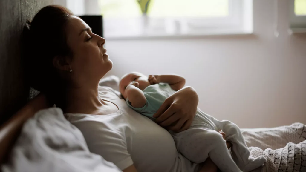 Woman experiencing loneliness in early motherhood lying down with newborn resting on her chest in a quiet bedroom.