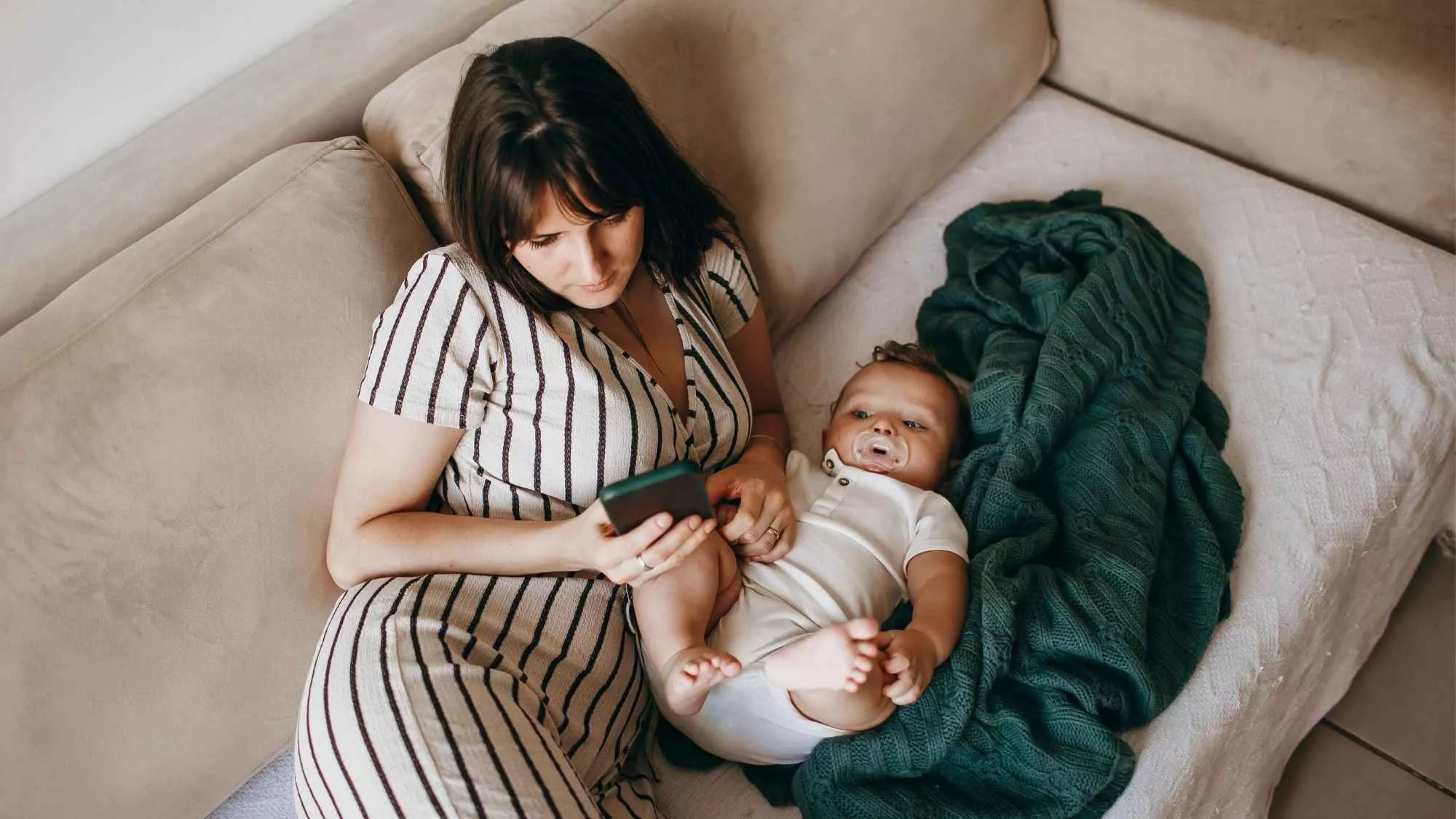 Mother sitting on couch with baby while looking at phone, showing emotional disconnection and maternal ambivalence in early motherhood.