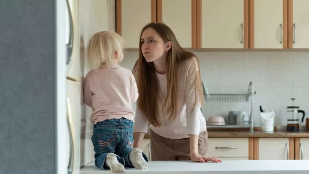 A mother feeling mom rage leans toward her toddler in a kitchen while the child kneels on the counter facing away.