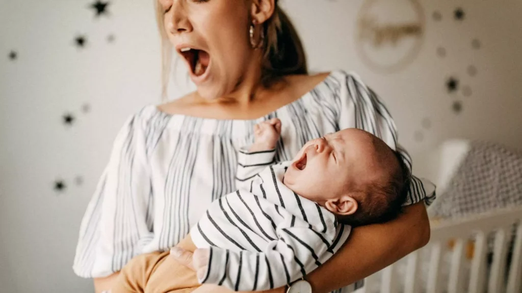A tired, but good enough mother yawns while holding a yawning baby in a nursery.