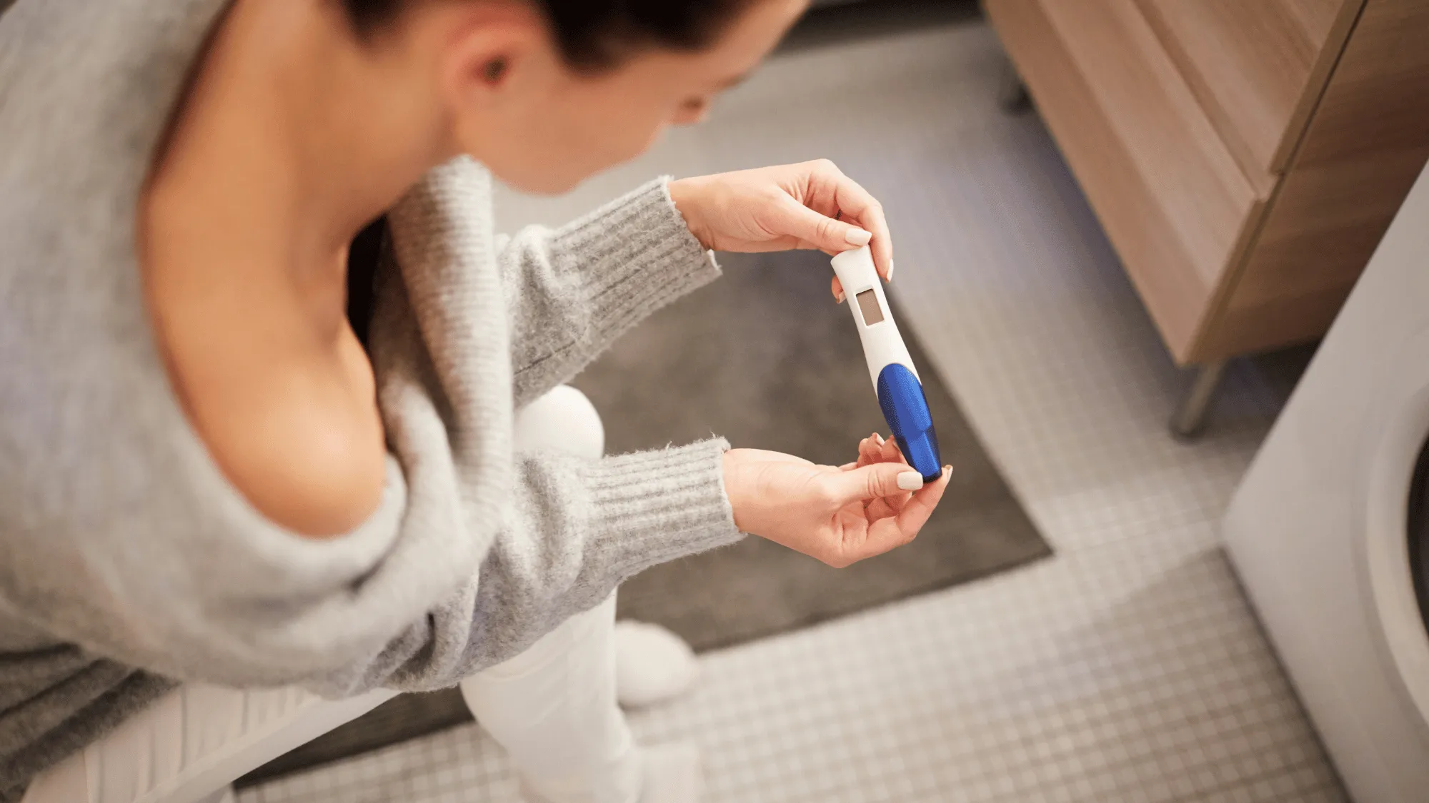 Woman sitting in a bathroom holding a pregnancy test and looking down anxiously, illustrating the intense fear and distress associated with tokophobia and pregnancy-related uncertainty.