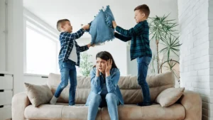 Two young boys play-fighting with pillows on a couch while their overwhelmed mother sits between them with her hands over her ears, illustrating how children's challenging behavior can stem from unmet needs rather than personal attacks on parents.