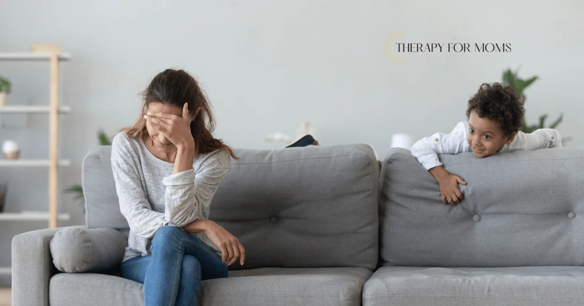 Stressed mother sitting on a couch with her hand covering her face while her child watches from behind the sofa, representing the emotional toll parents often feel when children act out or resist boundaries.